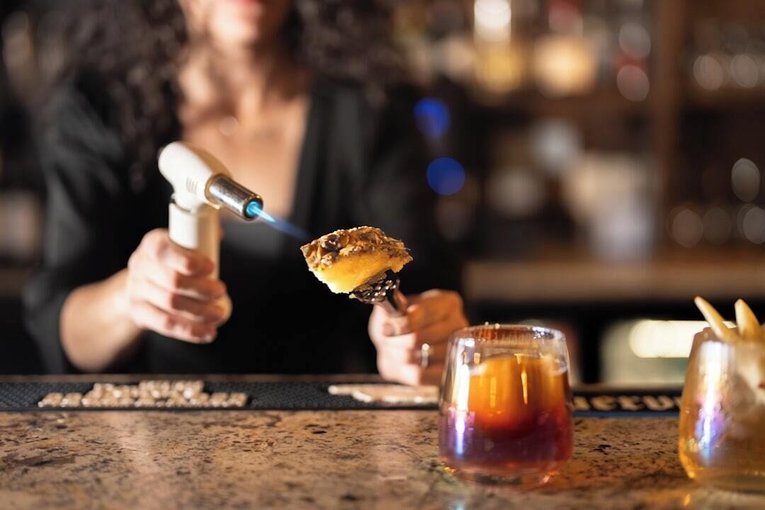 Iemand houdt een cocktailshaker met een blauwe vlam vast, samen met een glas met een rode drank en een fles met een gouden vloeistof.