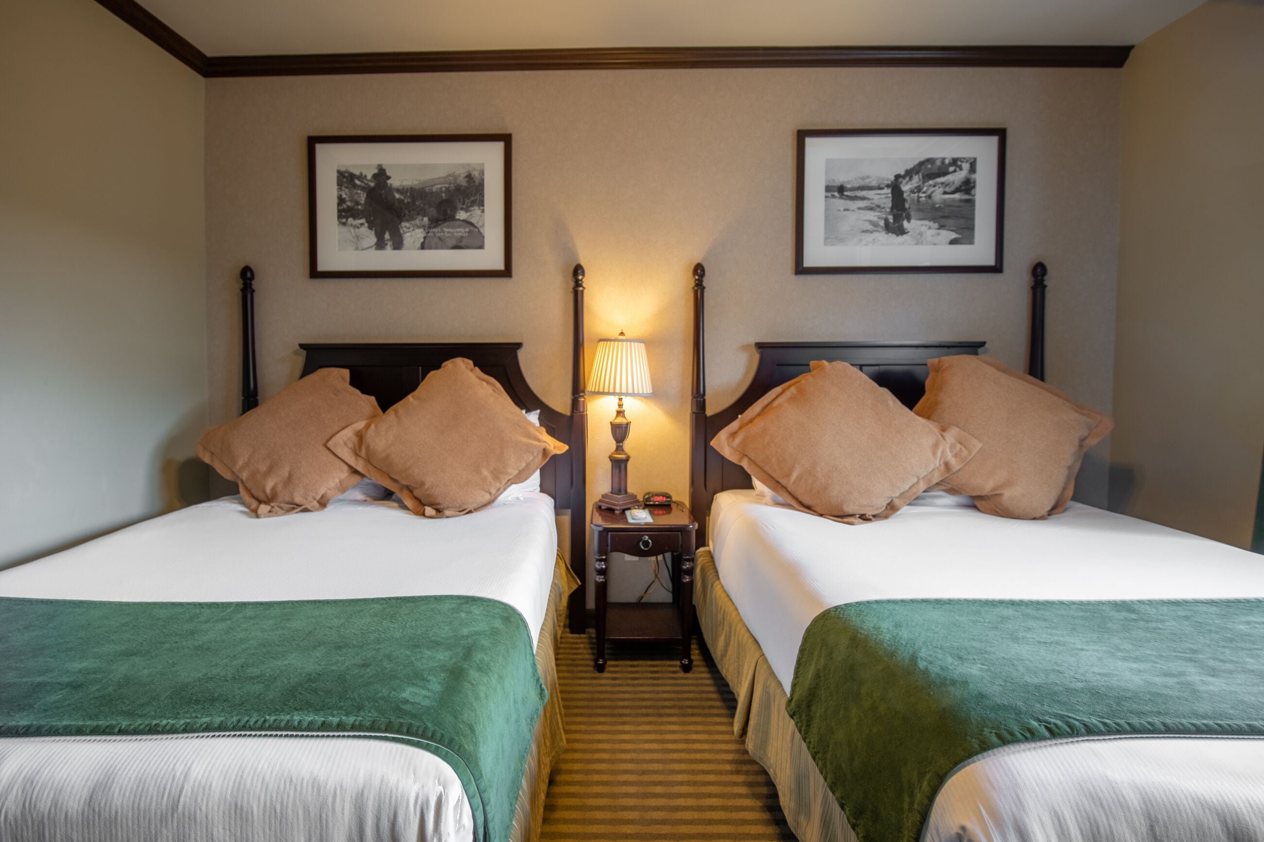 A hotel room with two beds, each with a green throw blanket, a nightstand with a lamp, and framed pictures on the wall.