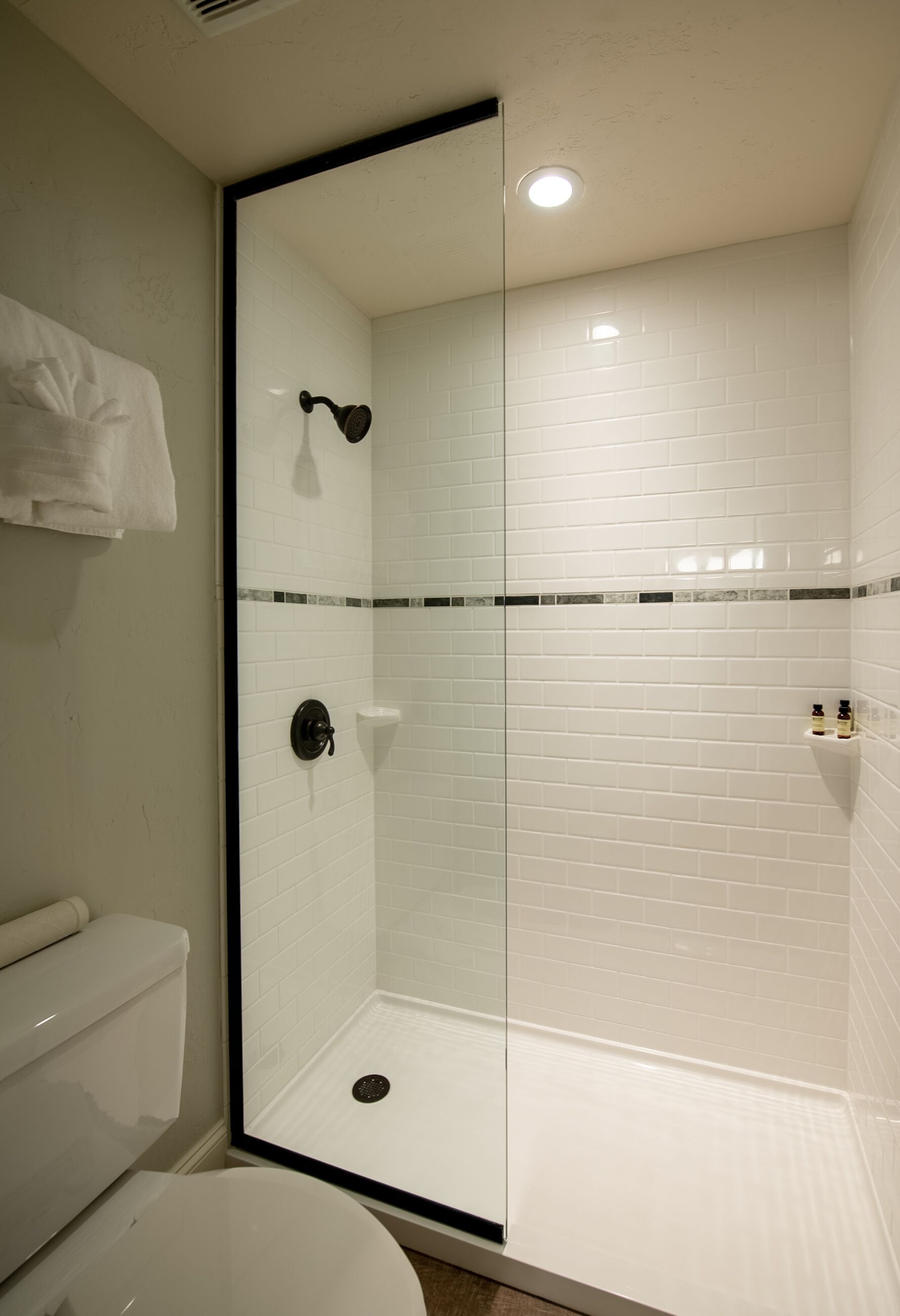Une salle de bain avec une douche à parois de verre, des murs recouverts de carreaux blancs, des toilettes et un porte-serviettes.
