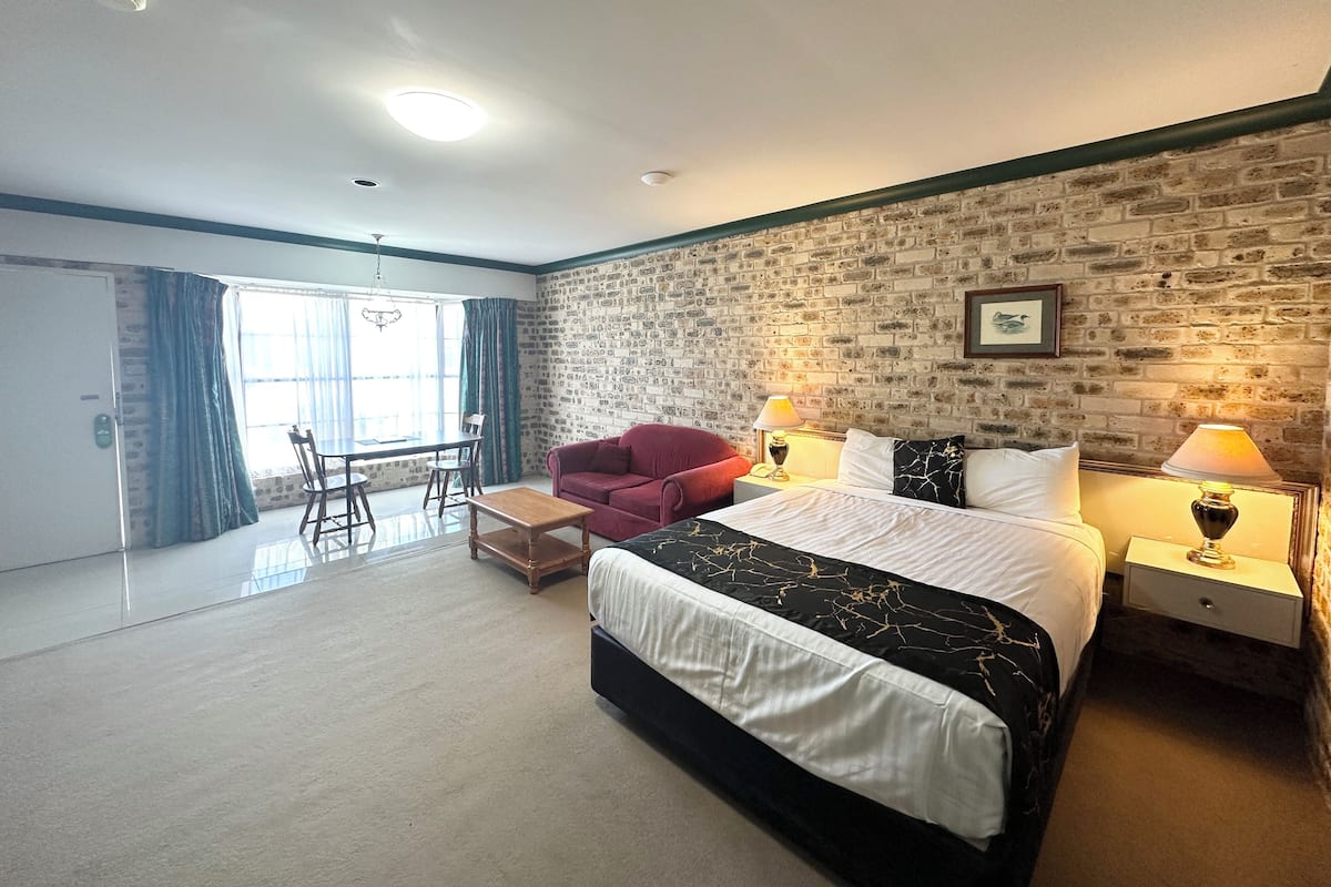 A hotel room with a brick wall, a large bed with white and black bedding, a red armchair, a wooden coffee table, and a desk with a lamp.