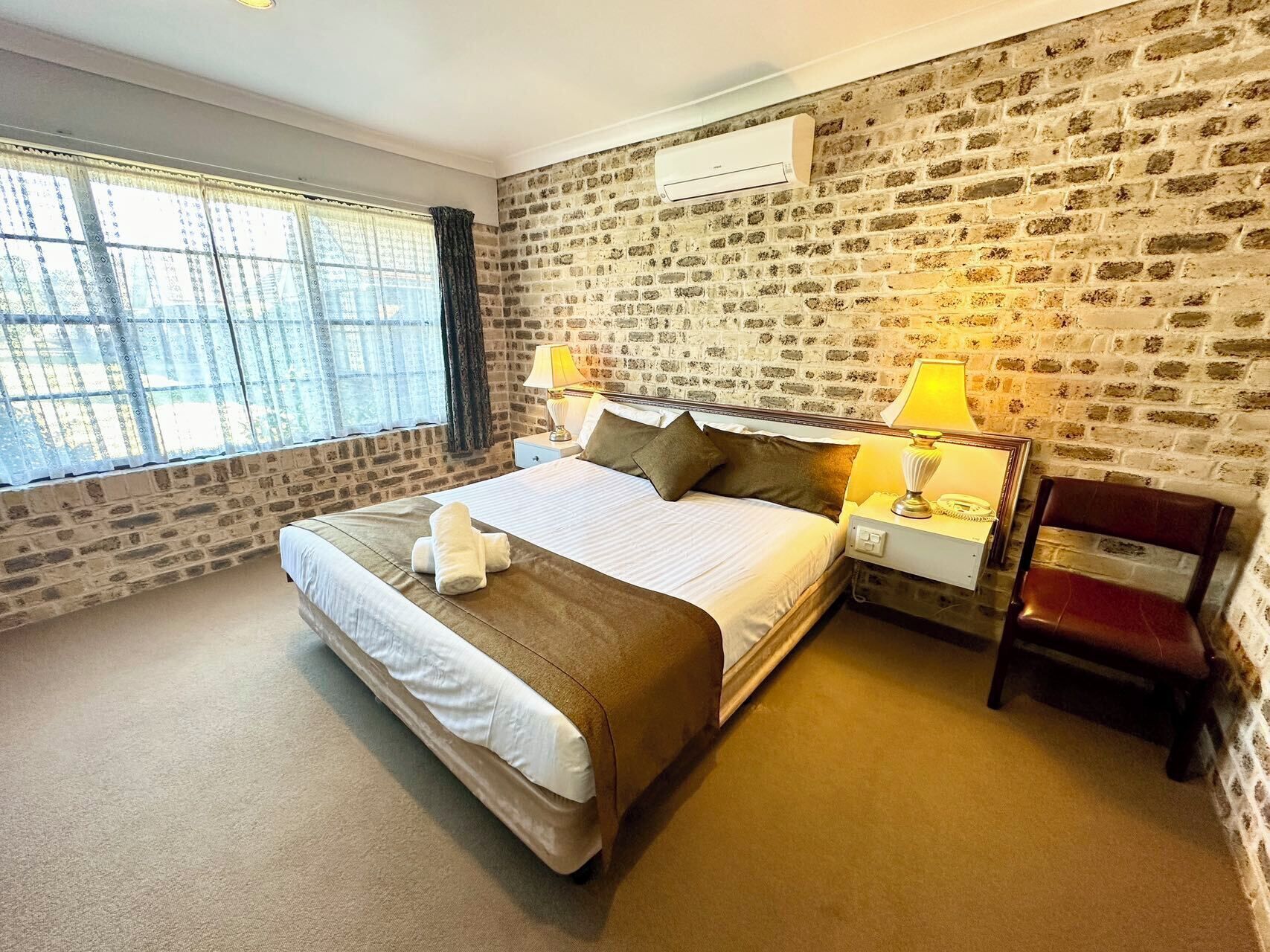 Habitación de hotel con una cama grande, dos mesitas de noche con lámparas, una pared de ladrillo y una ventana con cortinas.