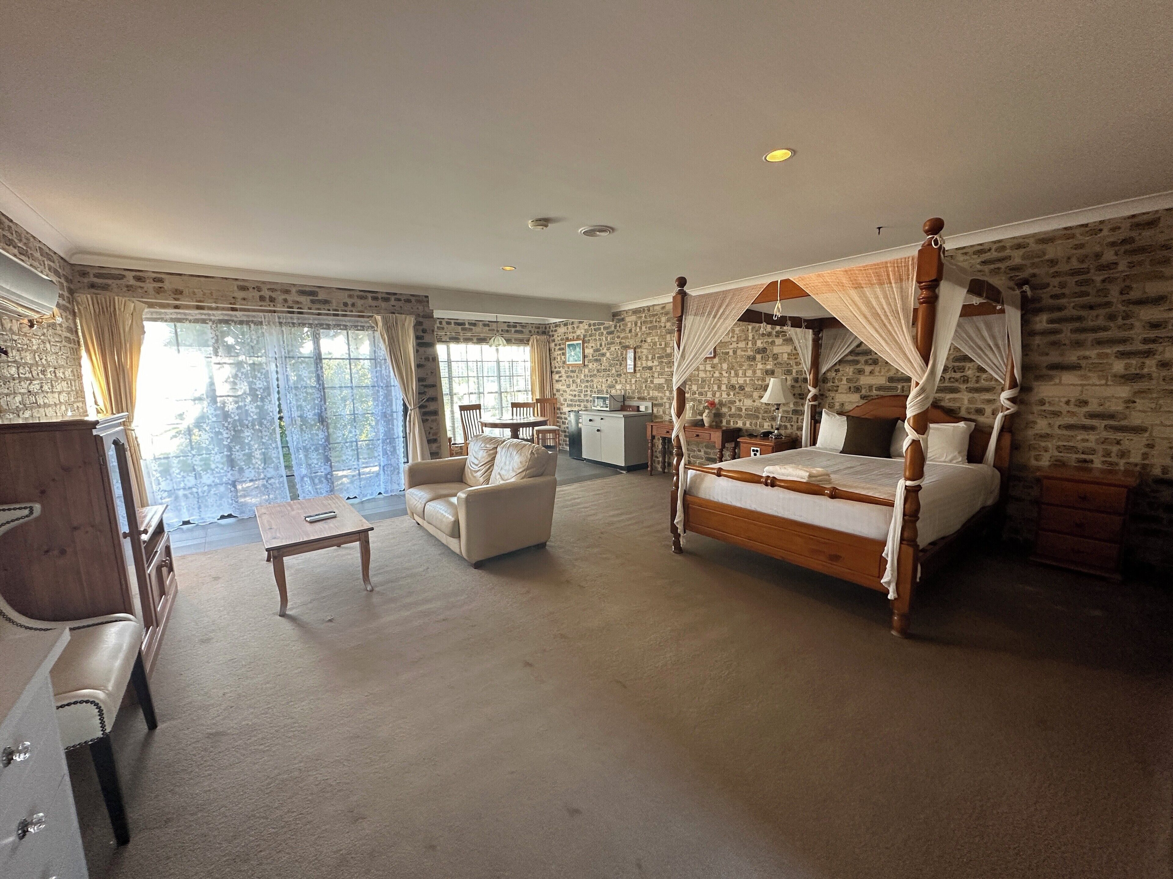 Un dormitorio espacioso con cama con dosel, una zona de estar con sofá y mesa de centro, y vistas al exterior a través de amplias ventanas.