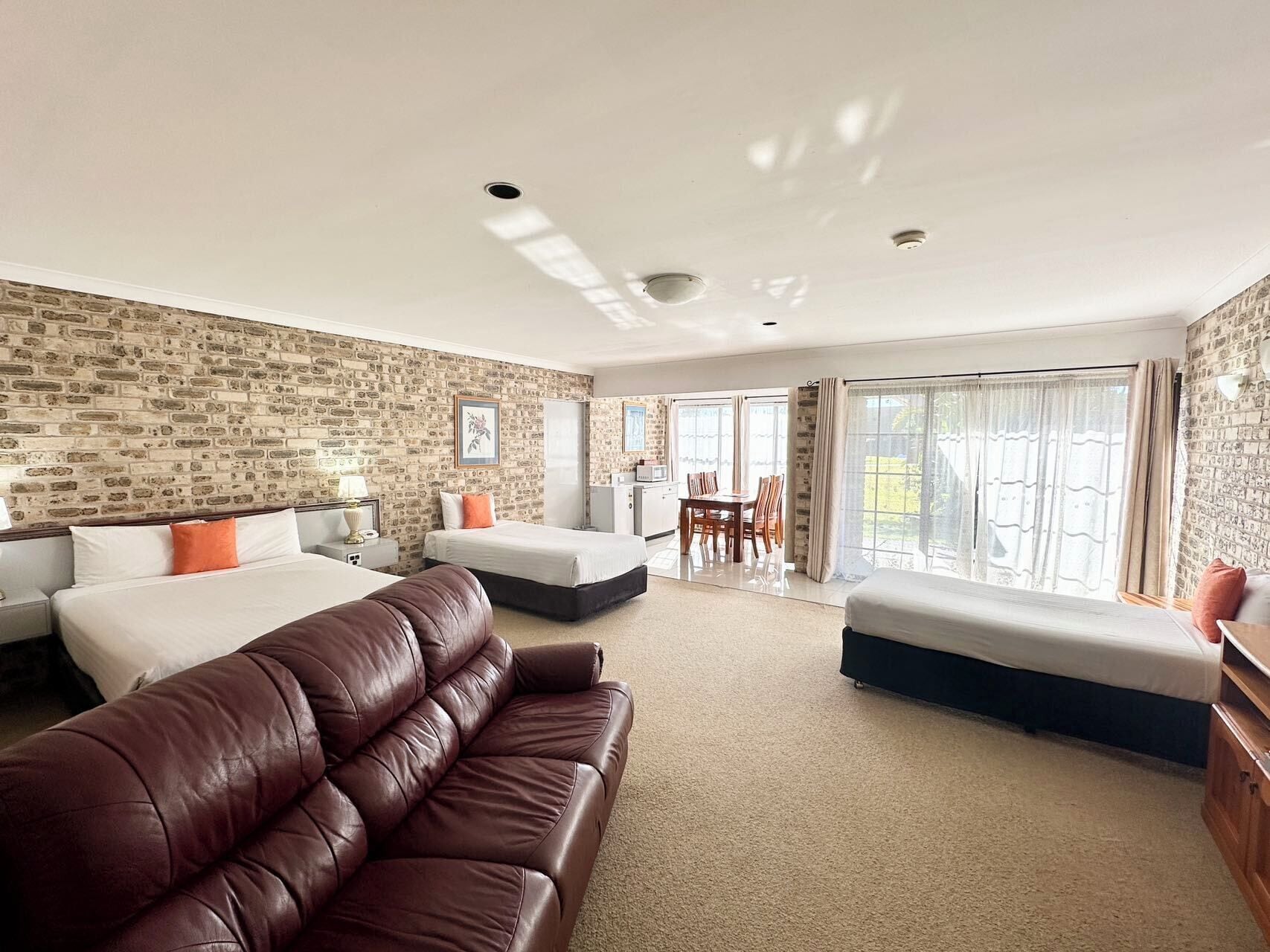 A hotel room with a brick-patterned wall, a large bed, a brown leather sofa, a dining table, and a sliding glass door leading to a balcony.