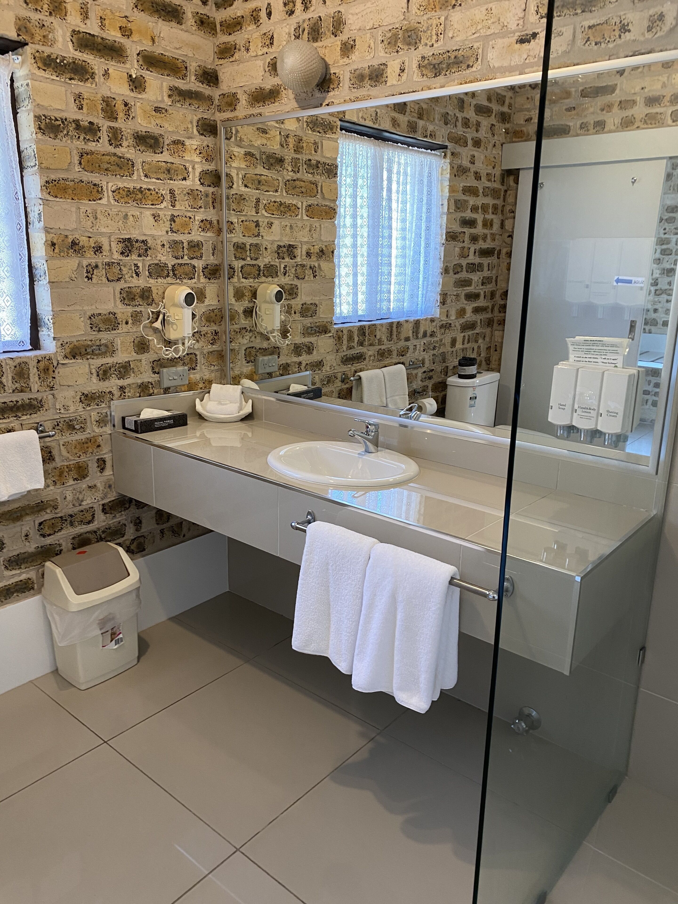 Ein modernes Badezimmer mit einer Wand aus Steinziegeln, einer Duschkabine aus Glas, einem großen Spiegel und einem weißen Waschbecken.