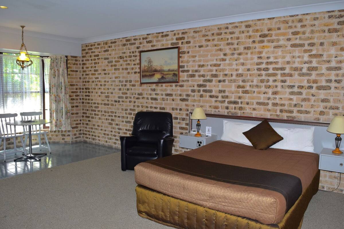 A hotel room with a brick wall, a bed with a brown headboard, a black leather chair, a small table, and a painting on the wall.