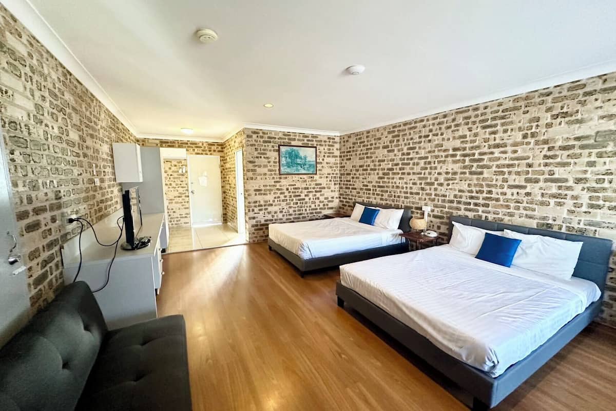 A hotel room with two beds, a desk, and a chair. The room has brick walls and wooden flooring.