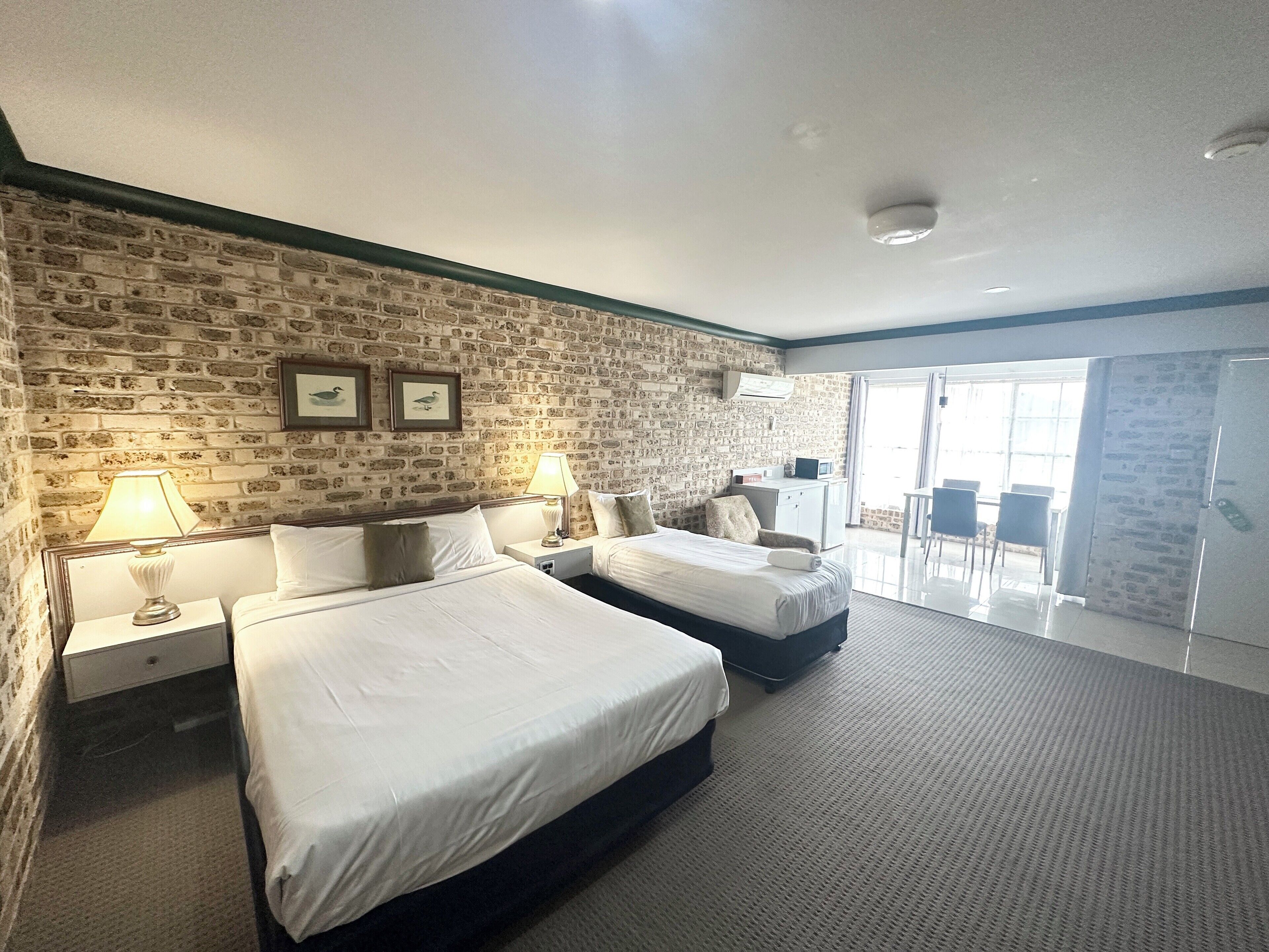 A hotel room with two beds, a brick-patterned wall, a desk with a computer, and a balcony with chairs.