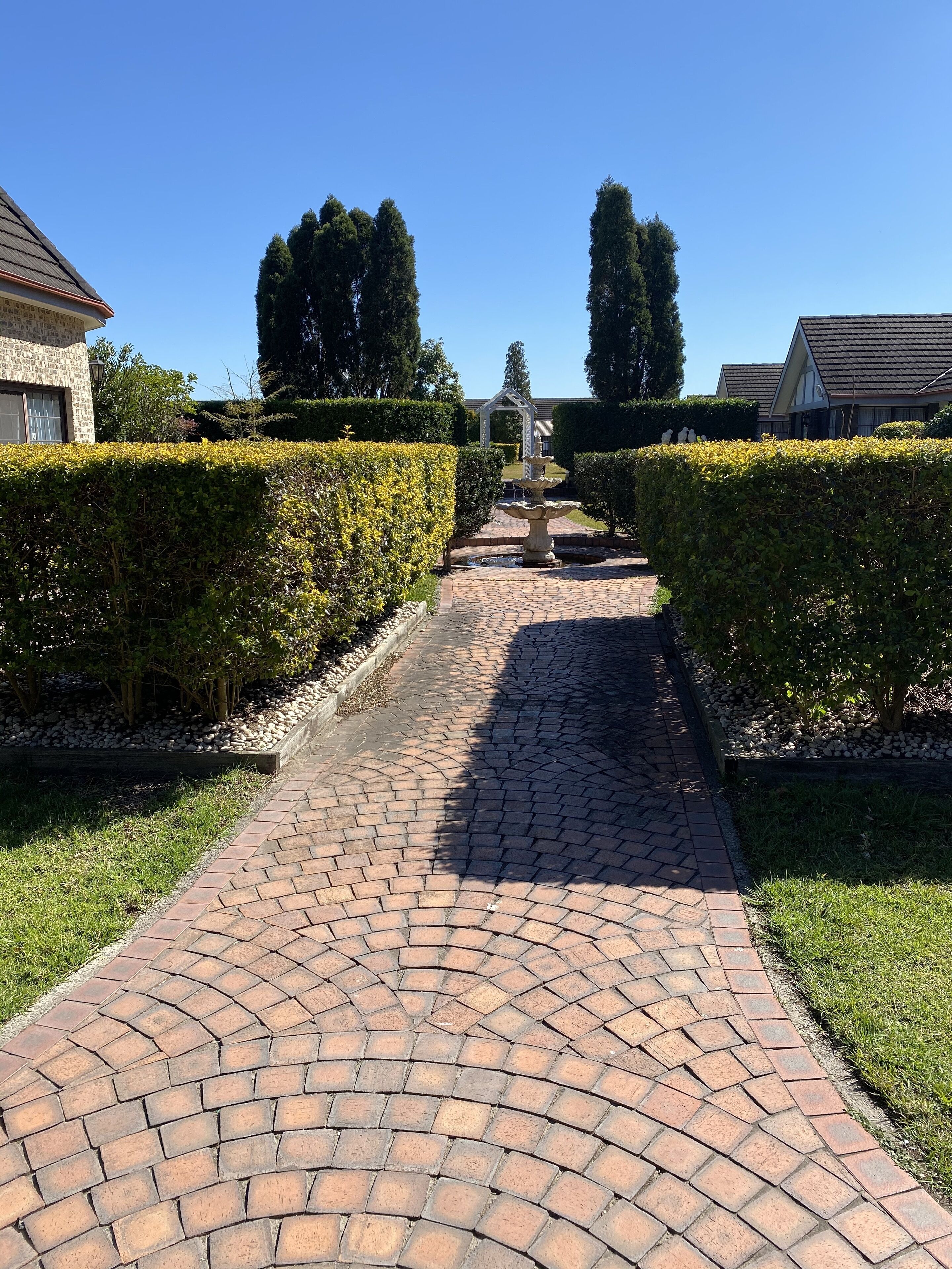 A paved pathway leading to a house with a stone facade, surrounded by well-maintained hedges and trees.