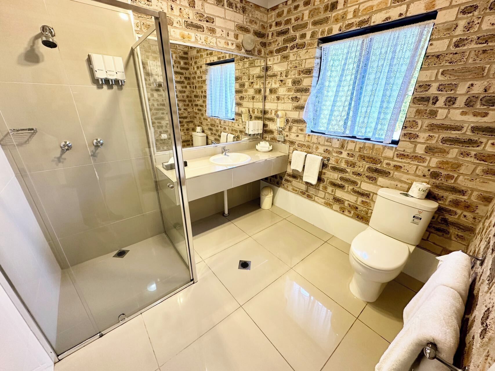 A bathroom with a glass shower enclosure, a white sink, a toilet, and a window with patterned curtains.