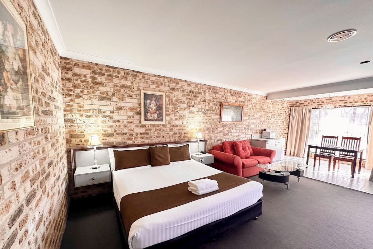 A hotel room with a brick wall, a large bed with white and brown bedding, a red sofa, a small table, and a chair.
