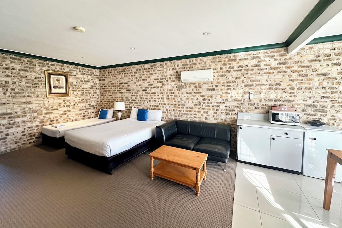 A hotel room with a brick-patterned wall, a bed with white linens, a black leather sofa, a wooden coffee table, a white desk with a microwave, and a framed picture on the wall.