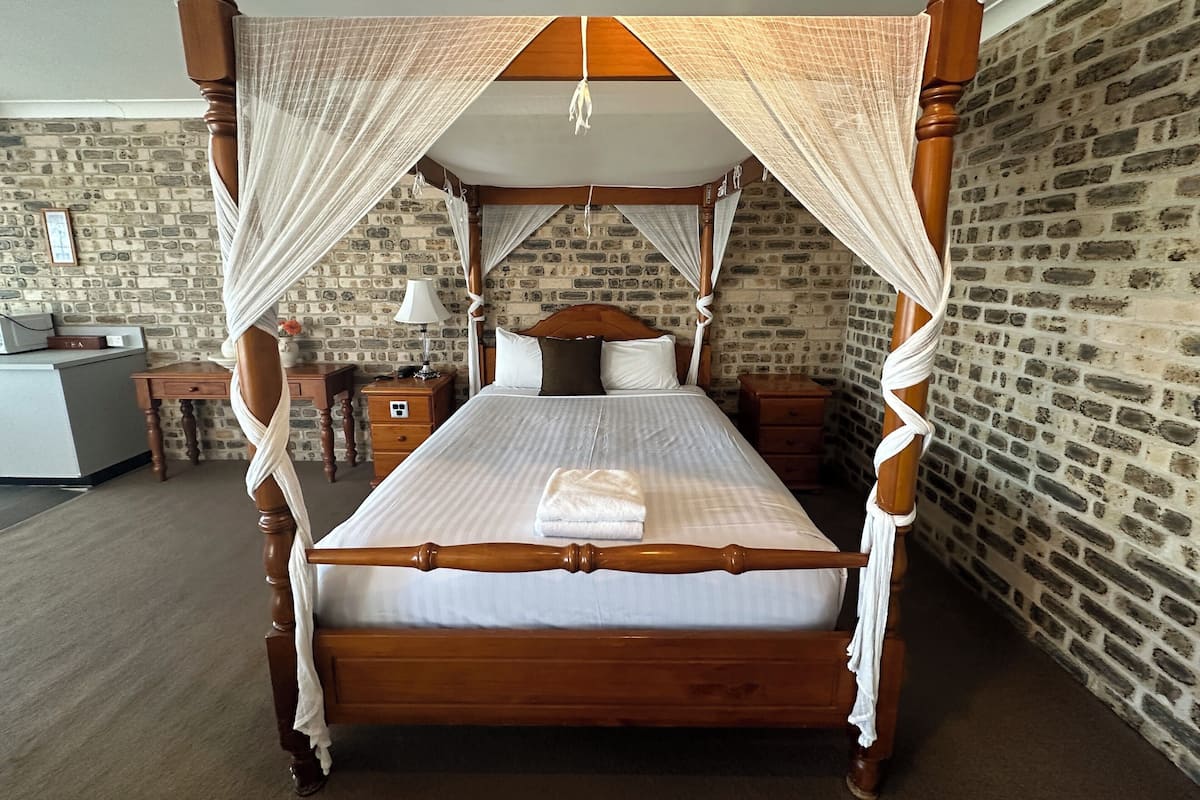 A four-poster bed with a canopy, a bedside table, a lamp, and a brick-patterned wall.