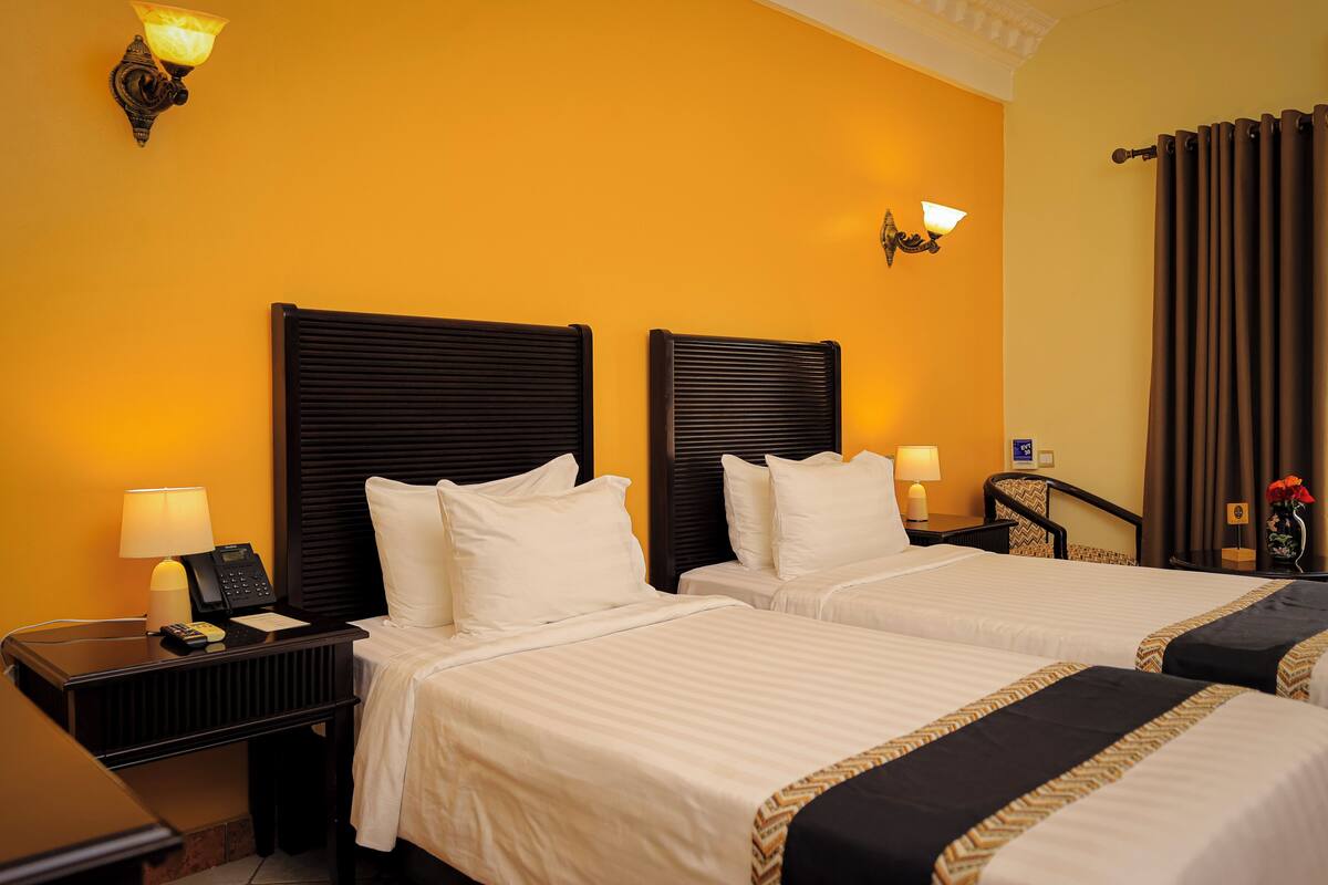 A hotel room with two beds, a nightstand with a lamp, a telephone, and a small table with a vase of flowers.