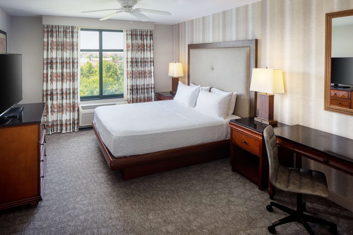 A hotel room with a large bed, two bedside tables with lamps, a desk with a chair, a television, and a window with curtains.