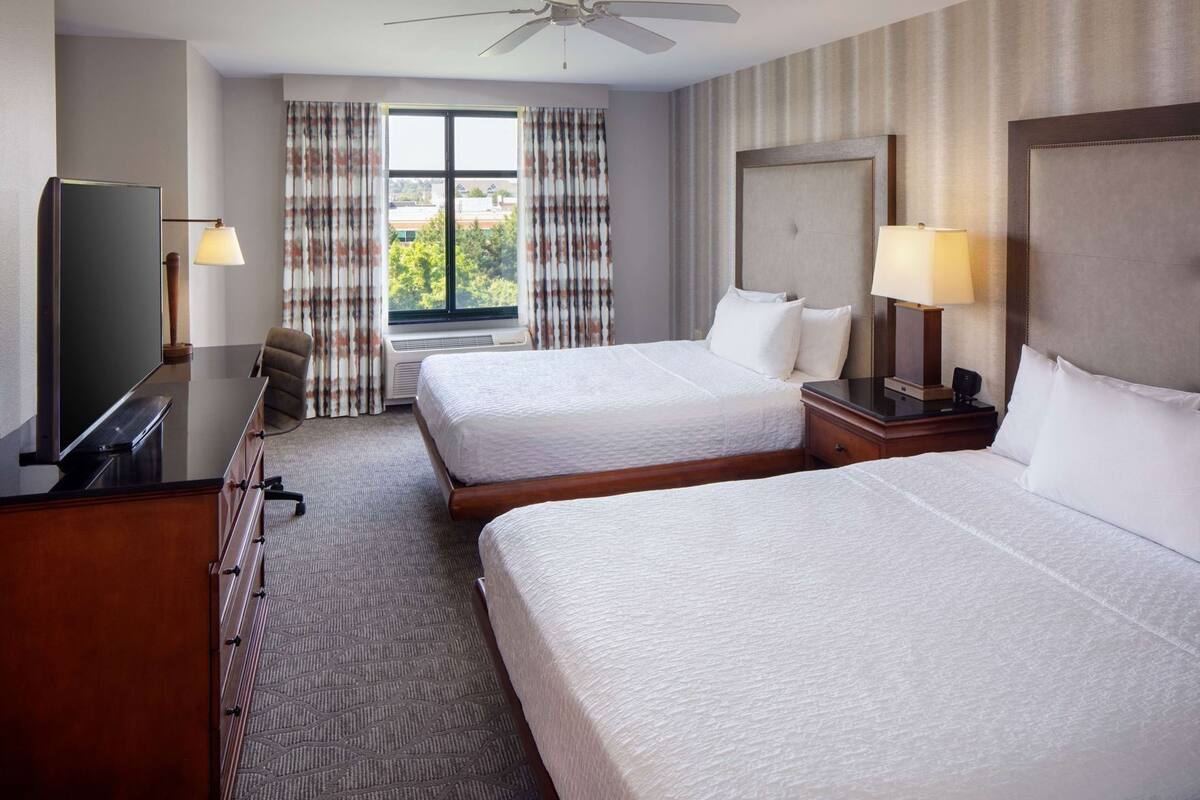 A hotel room with two beds, a desk with a television, a ceiling fan, and a window with curtains.