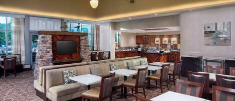 A spacious hotel lobby with a stone fireplace, comfortable seating, and a modern kitchen area.