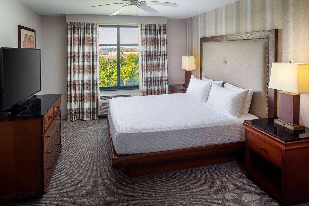 A hotel room with a large bed, a TV, a desk, and a window with curtains.
