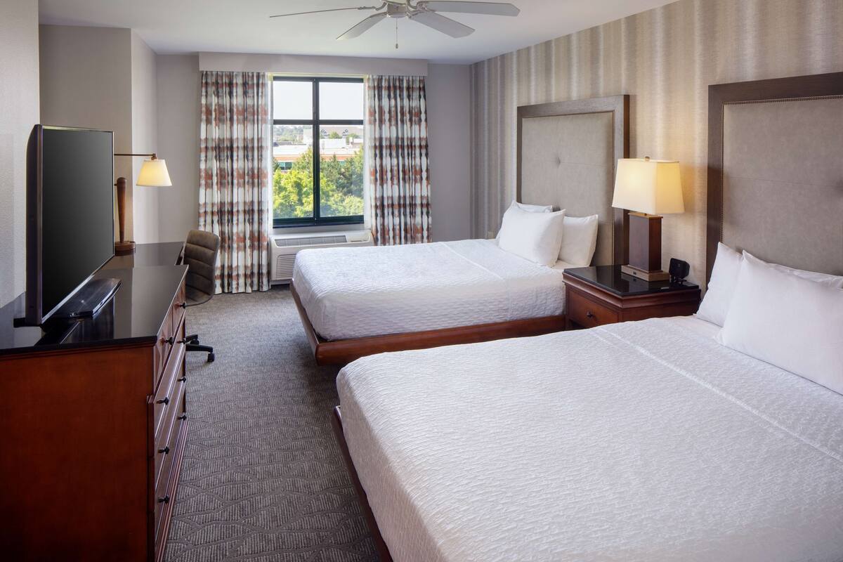 A hotel room with two beds, a desk with a TV, a window with curtains, and a ceiling fan.