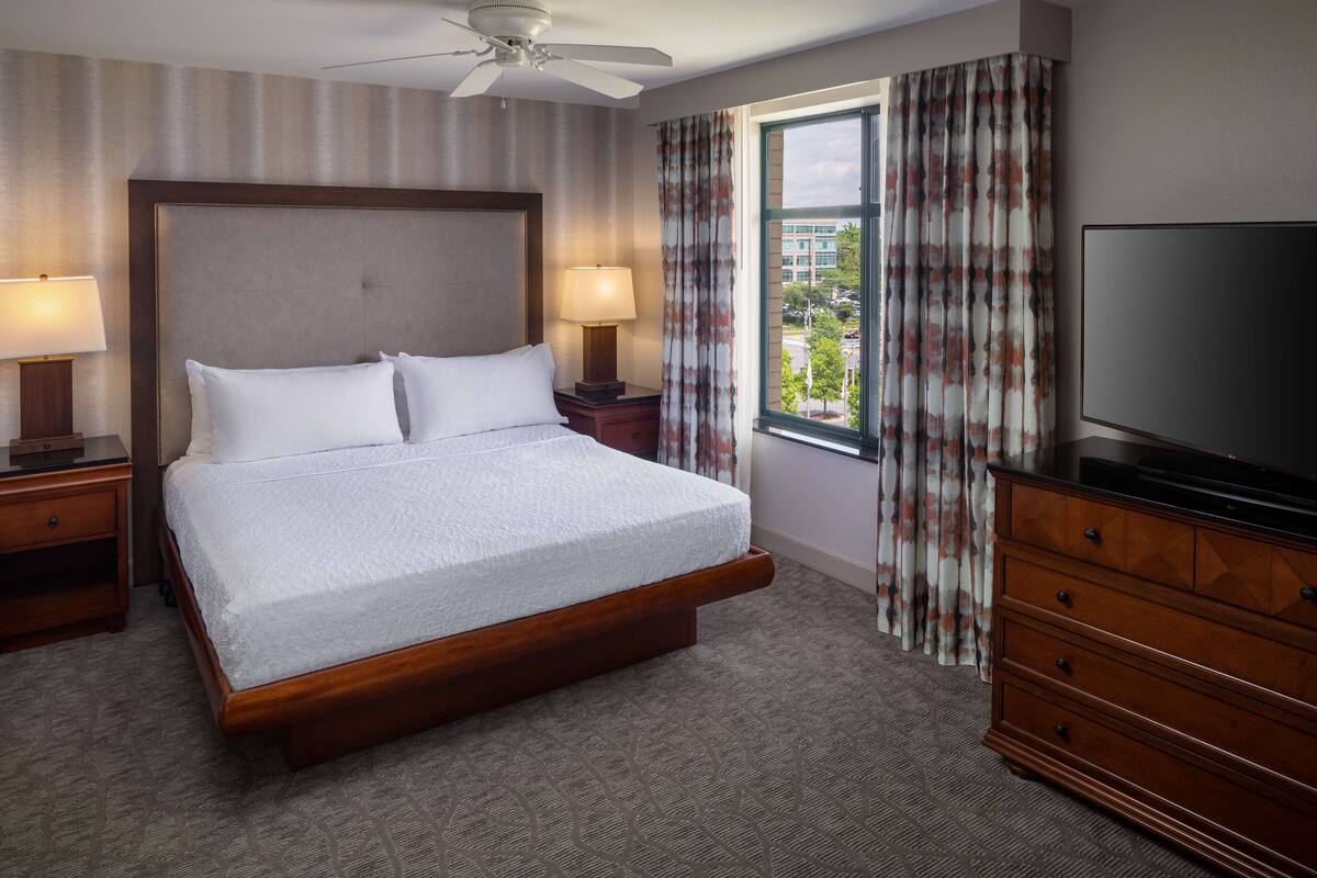 A hotel room with a large bed, two bedside lamps, a TV, a dresser, and a window with curtains.