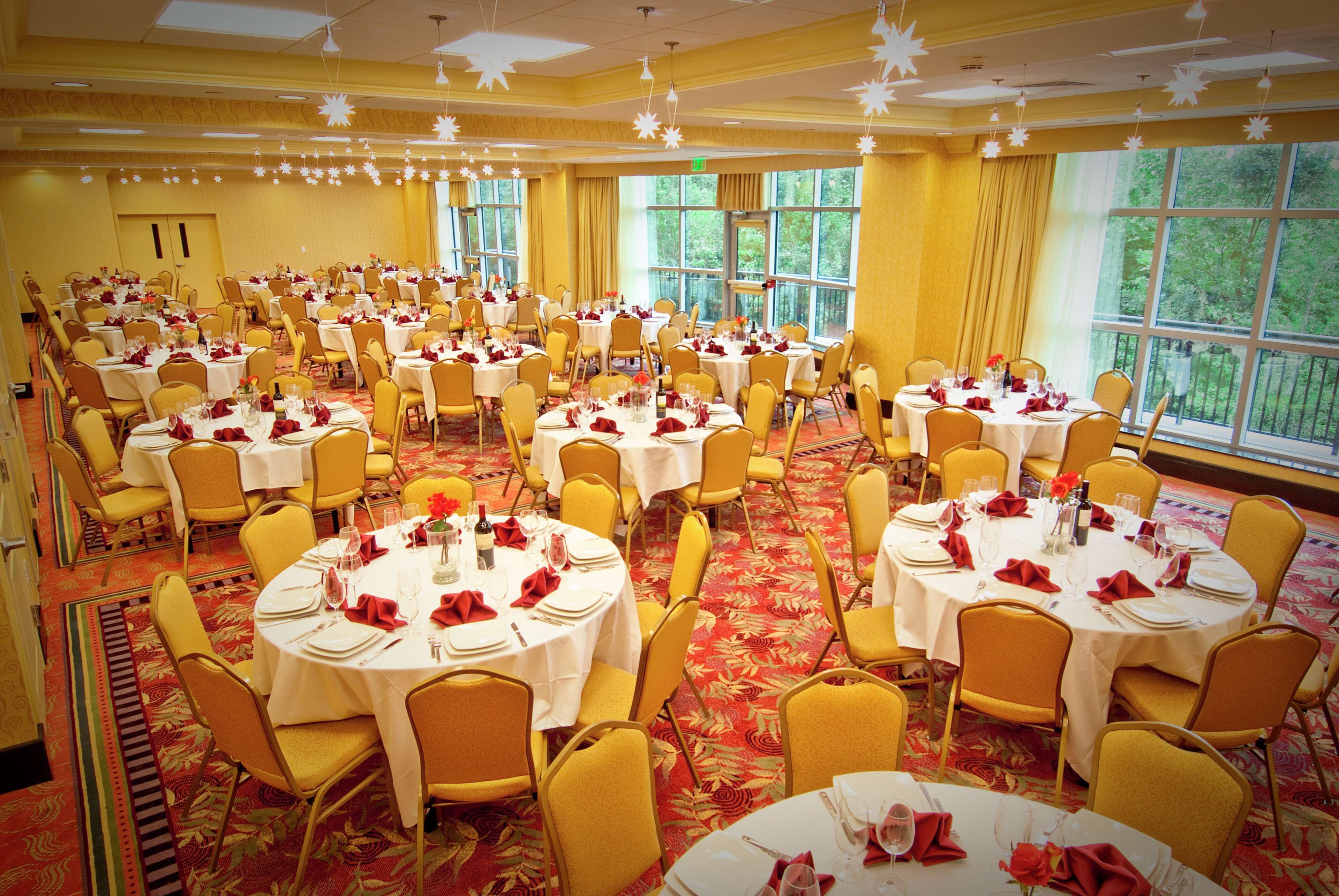 Un salón de banquetes con varias mesas redondas dispuestas para comer, sillas amarillas y grandes ventanales que ofrecen vistas al exterior.
