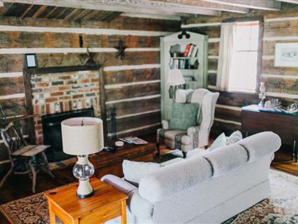 A rustic log cabin interior with a fireplace, a green armchair, a white sofa, and a wooden coffee table.