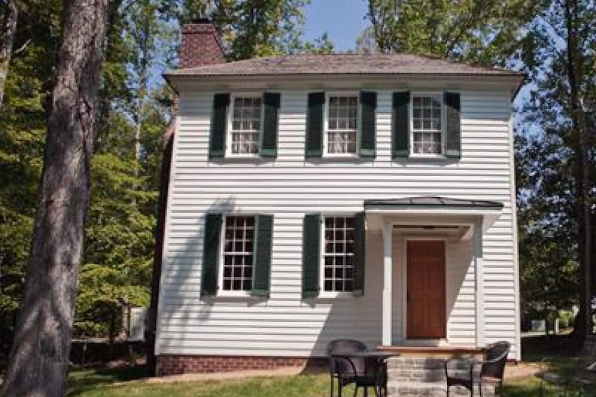 A two-story white house with green shutters, a front door, and a small patio with two chairs.
