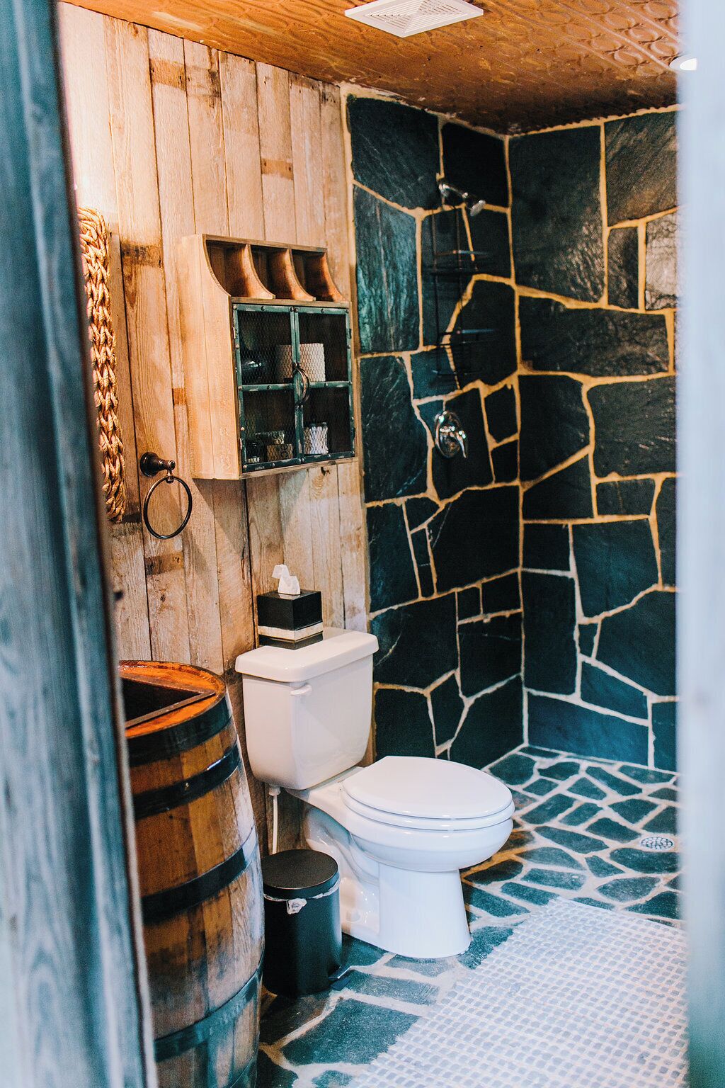 Un baño rústico con pared de piedra, paneles de madera, un inodoro y un barril.