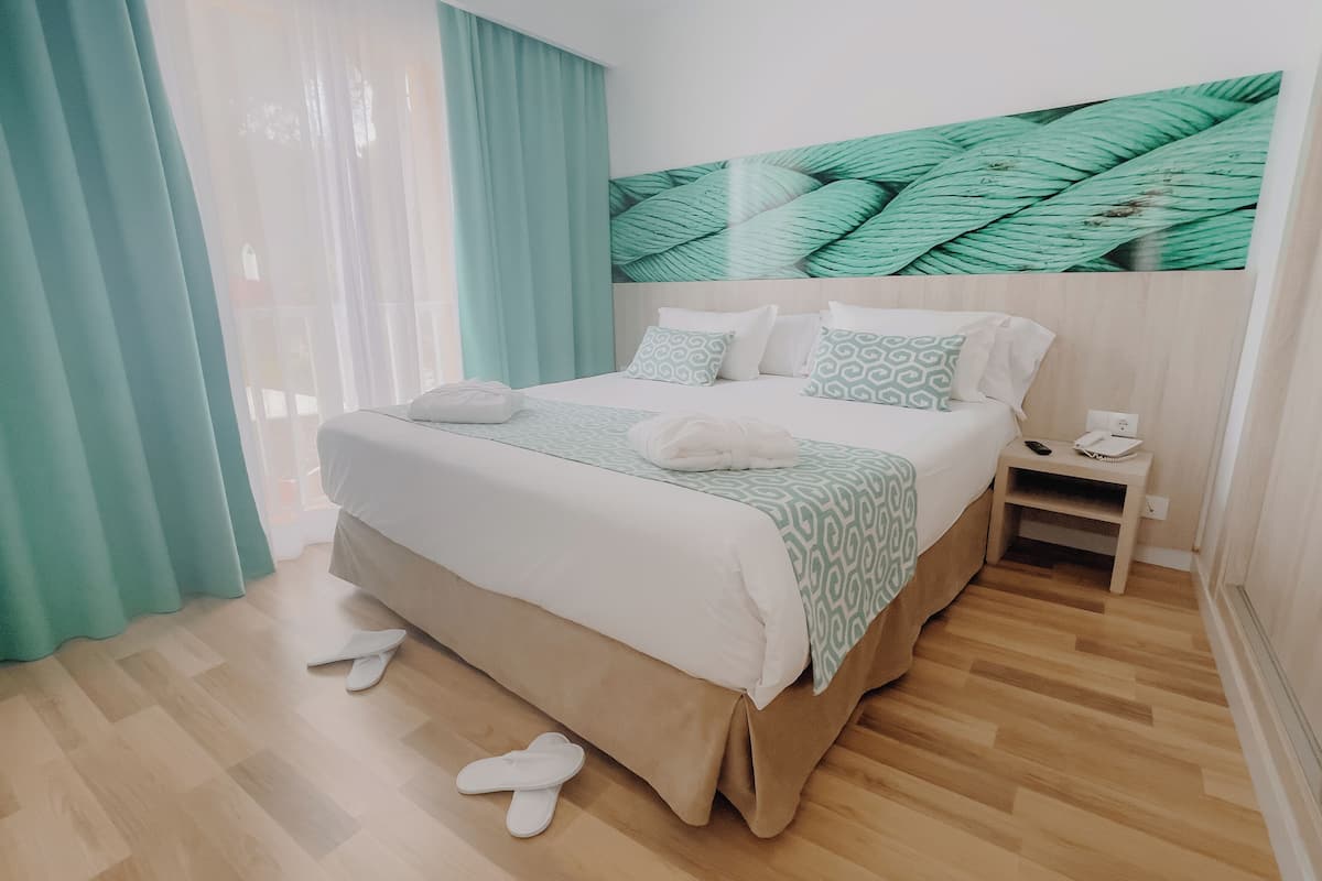 Una cama bien hecha con una colcha estampada, un par de zapatillas en el suelo y una mesita de noche con un teléfono y un control remoto.