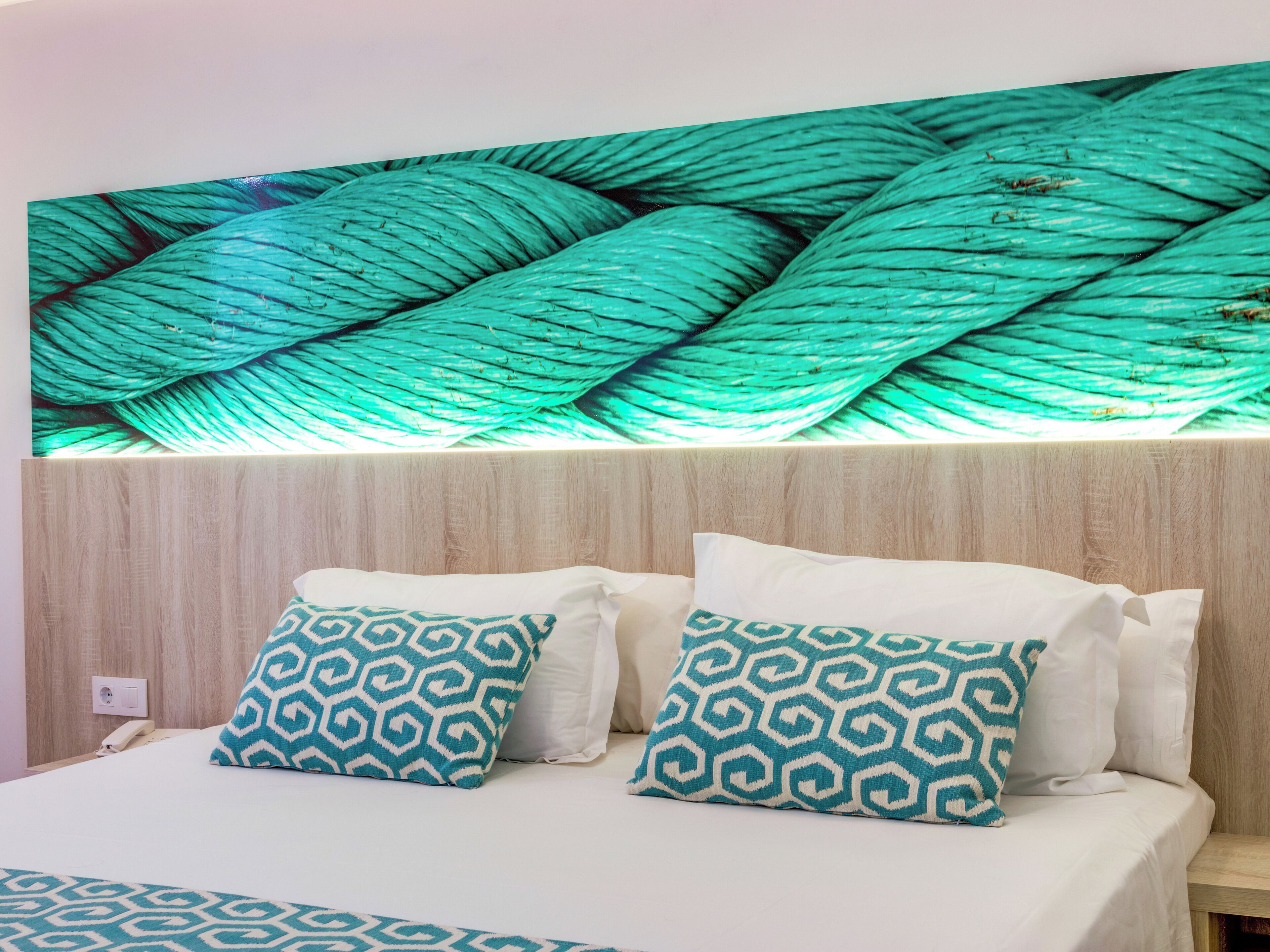 Une chambre d’hôtel avec un lit, deux coussins à motifs, une lampe fixée au mur et une prise de courant.