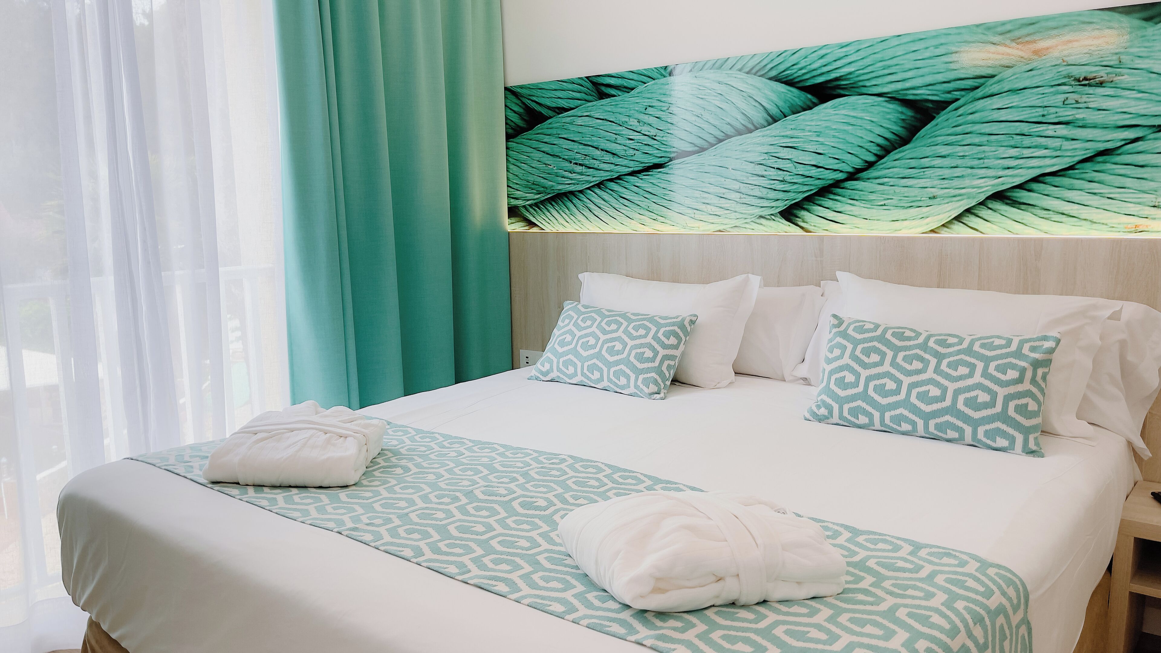 Een net opgemaakt bed met gestreepte kussens en een handdoek, een houten hoofdbord, en uitzicht op een groen gordijn.