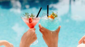 Two hands holding cocktail glasses with straws, one with a strawberry garnish and the other with an orange slice, over a pool.