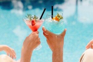 Dos manos sosteniendo copas de cóctel con pajitas, una con una fresa y la otra con una rodaja de naranja, sobre una piscina.