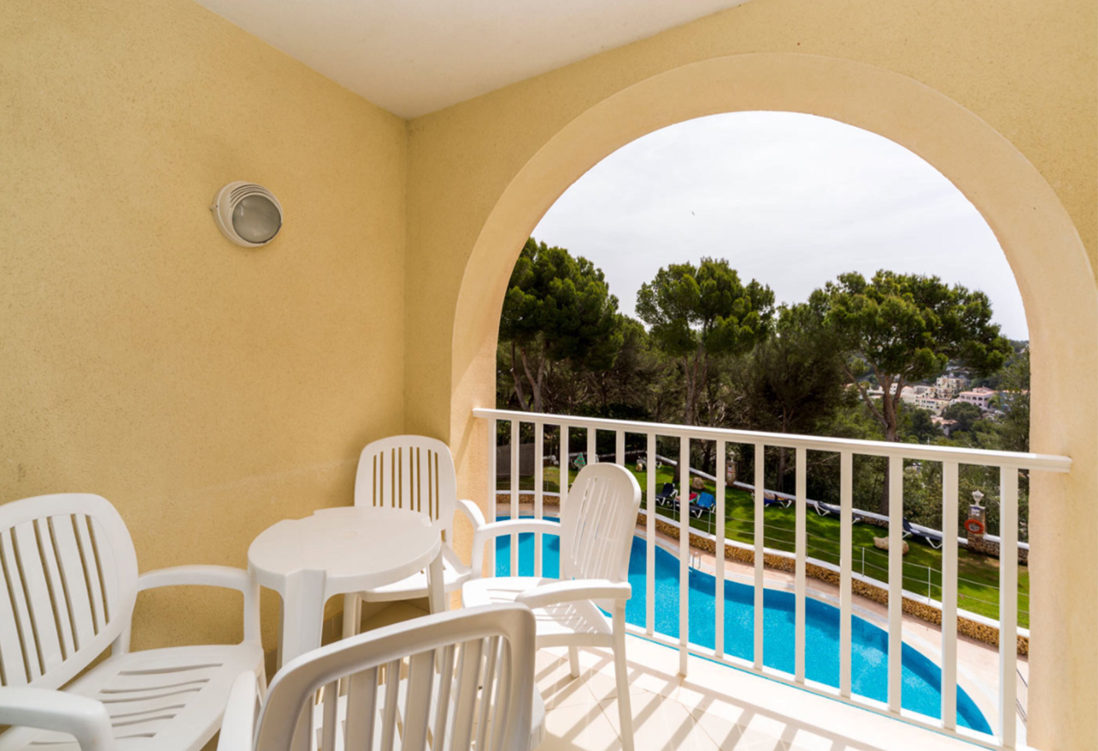 Un balcón con sillas de plástico blanco y una mesa pequeña, con vista a una piscina y un jardín ajardinado.