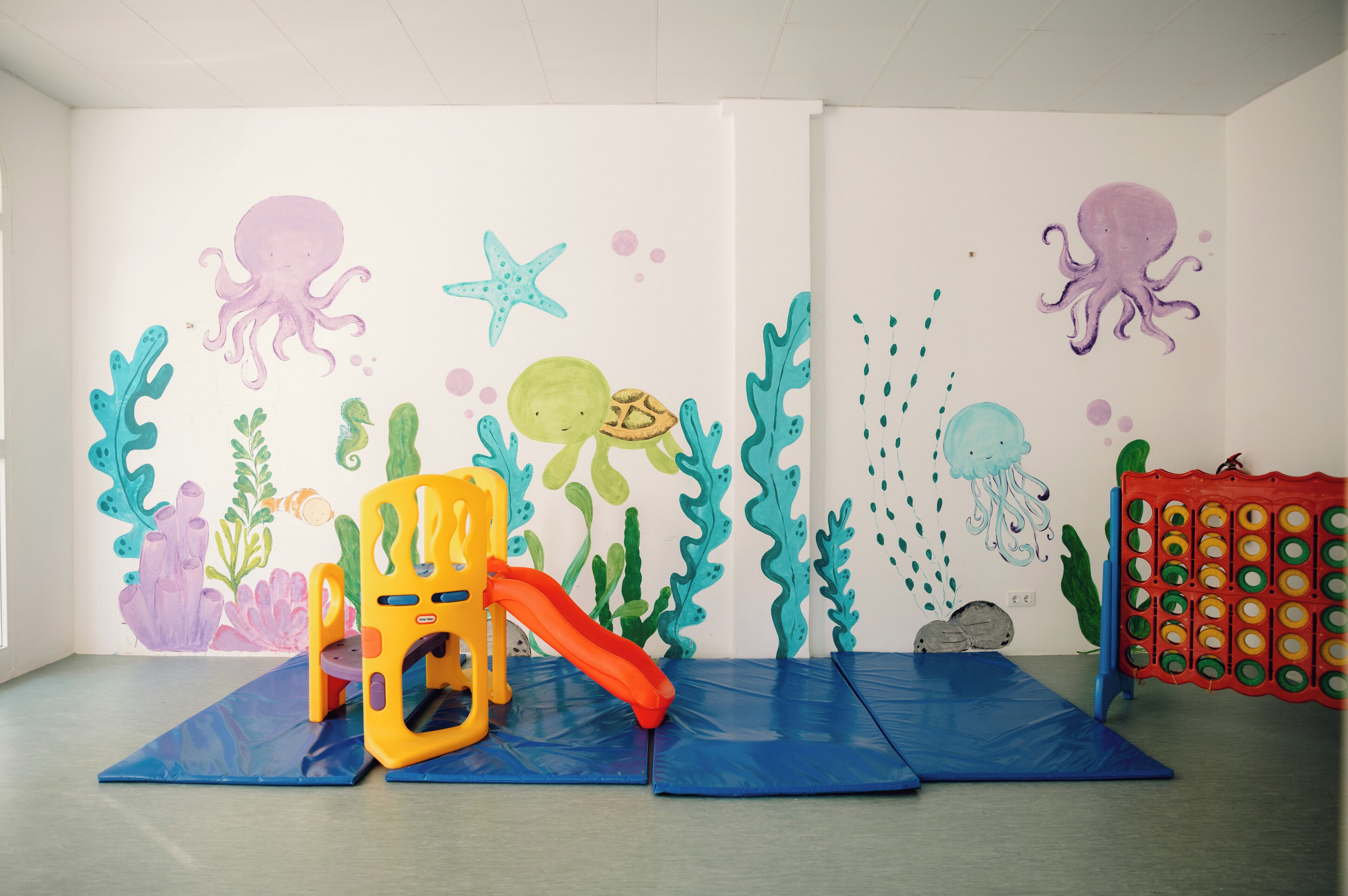 Une pièce dotée d’un espace de jeux comprenant un toboggan, une chaise jaune et une fresque murale colorée représentant des créatures marines.