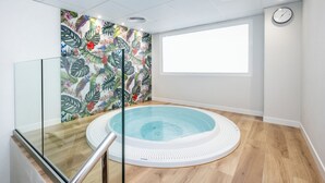 Una piscina interior moderna con un mural de hojas tropicales, barandilla de vidrio y suelo de madera.