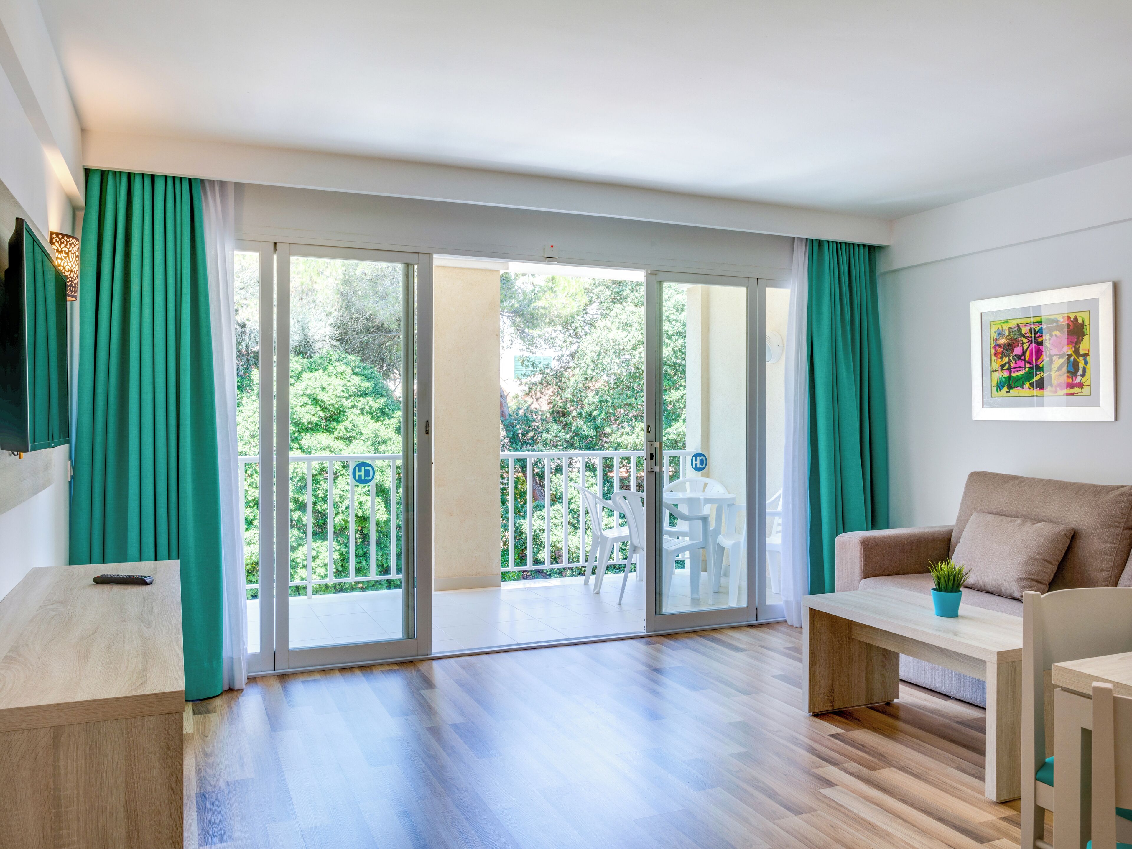 Una sala de estar moderna con un sofá, una mesa de centro y un televisor. Hay una puerta corrediza de vidrio que da a un balcón con mobiliario exterior.