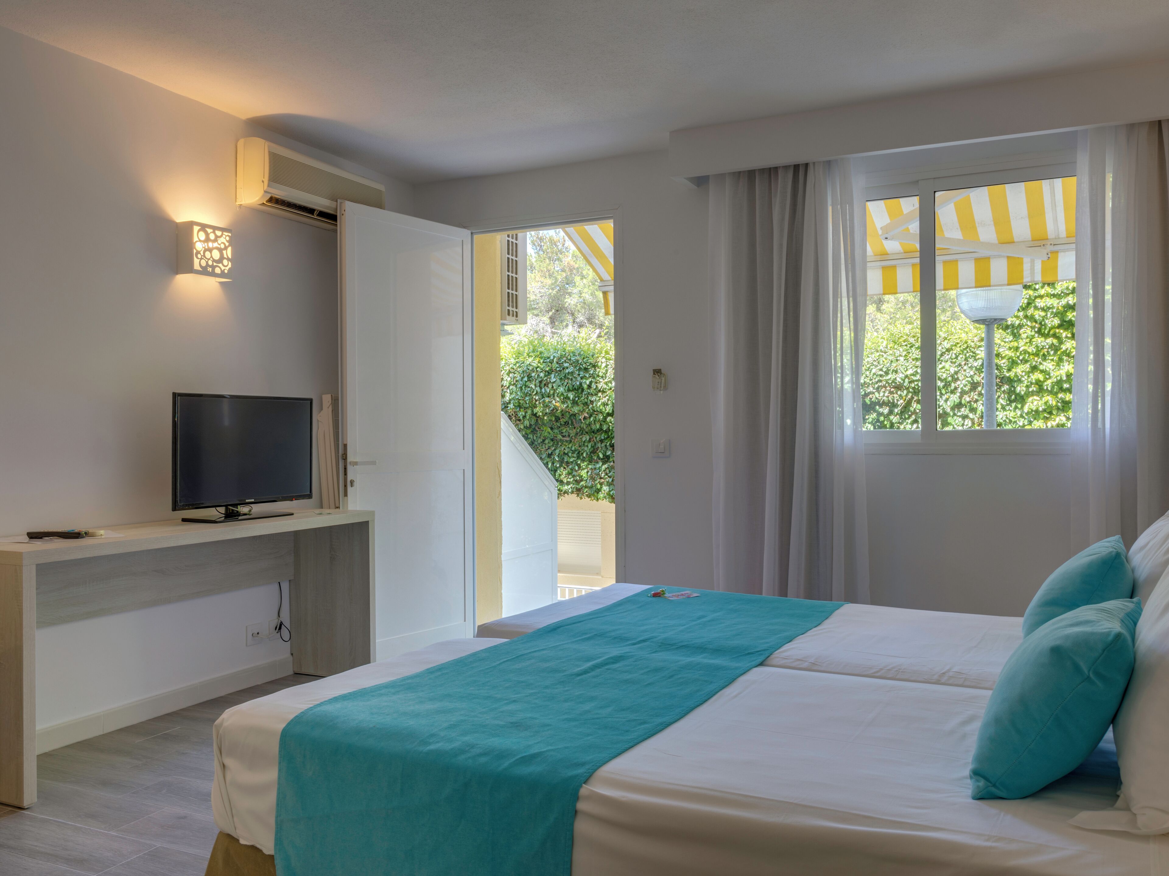 Un dormitorio con cama, televisor sobre un mueble bajo, ventana con cortinas y vista a zonas verdes.