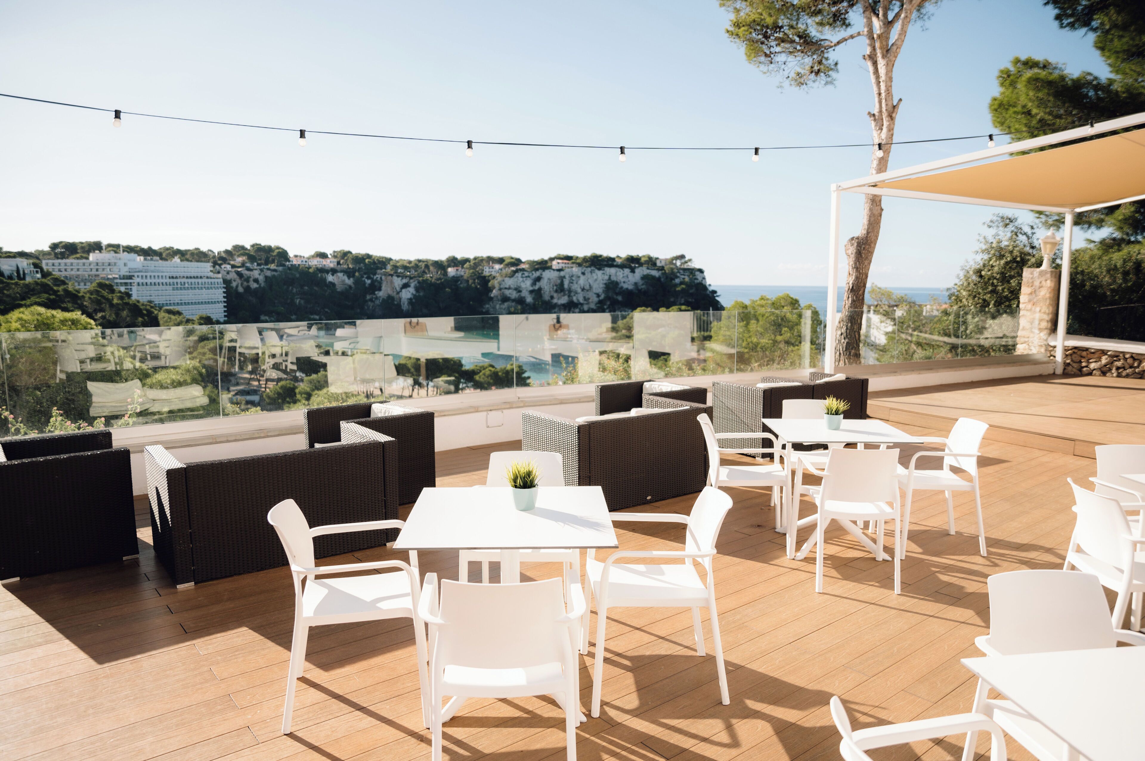 An outdoor patio with white tables and chairs, a pergola with a beige canopy, and a view of a coastal landscape.