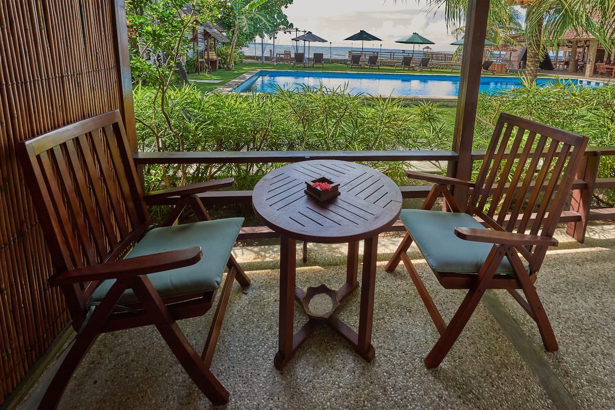 Terraza de madera con dos sillas y una mesa, con vistas a una piscina y abundante vegetación.
