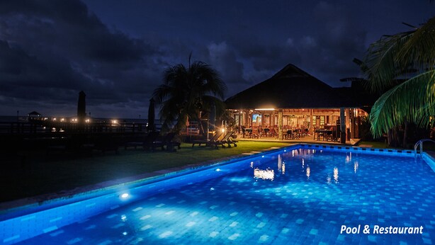 Zona de piscina y restaurante de noche, con piscina iluminada, palmeras y un edificio con techo de paja.