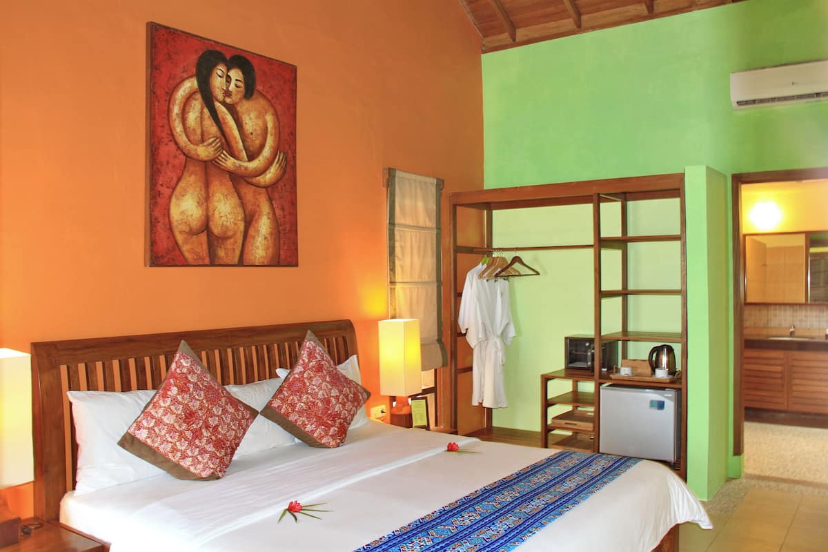 Habitación de hotel con cama, almohadas, un cuadro, una mesita de noche, lámpara, bata, refrigerador minibar y vista al baño.
