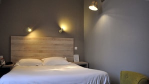 Una habitación de hotel con cabecera de madera, cama con ropa blanca, mesita de noche, lámpara y un sillón verde.