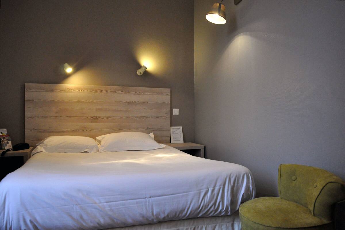 Quarto de hotel com cabeceira de madeira, cama com roupa de cama branca, mesa de cabeceira, luminária e poltrona verde.