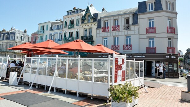 Filas de mesas de comedor al aire libre con sombrillas rojas, un letrero de bar y restaurante, y un letrero de hotel.