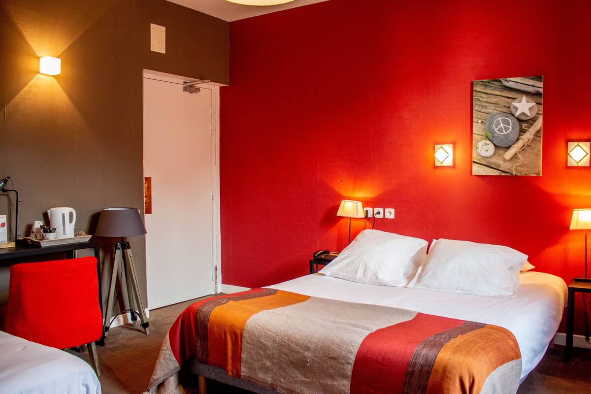 Quarto de hotel com uma cama, duas luminárias de cabeceira, uma escrivaninha com luminária, uma cadeira e uma obra de arte na parede.