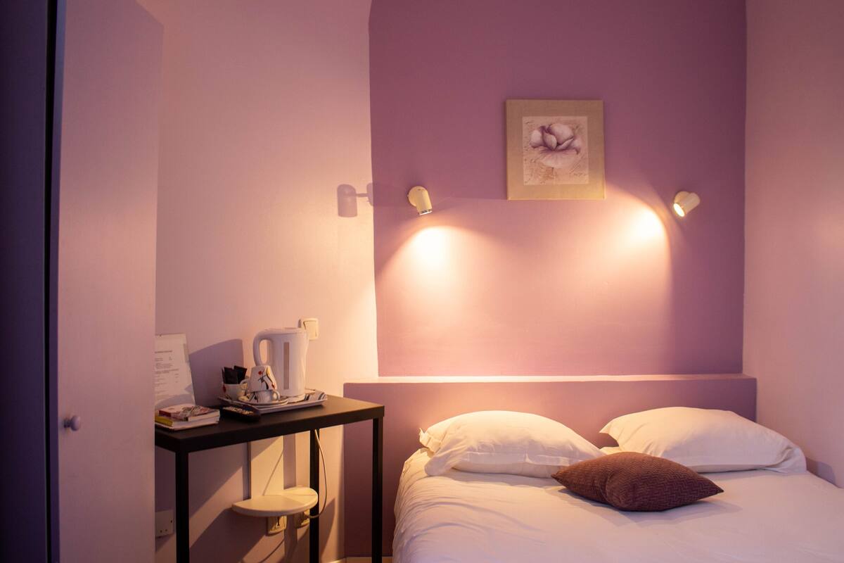 Quarto de hotel com cama, almofadas, mesa de cabeceira com chaleira, chávena e comando. Aparelho de iluminação fixado na parede e quadro enquadrado.