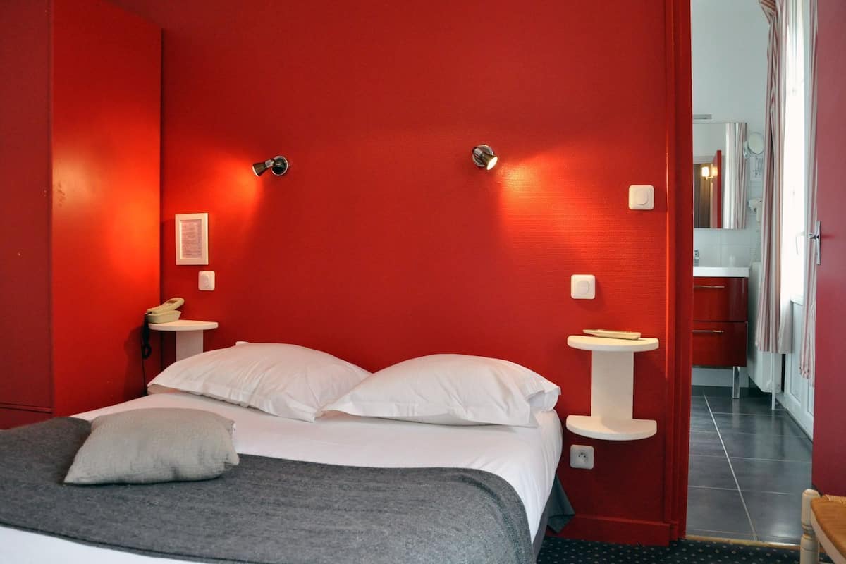 Quarto de hotel com parede vermelha, cama com roupa de cama branca, mesa de cabeceira com telefone e pia pequena branca.