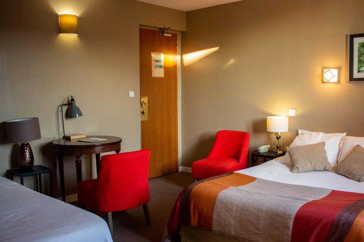 Quarto de hotel com cama, duas cadeiras, escrivaninha e mesa de cabeceira.