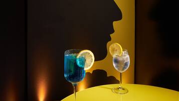 Une table jaune sur laquelle reposent deux verres contenant des boissons bleues et incolores, un magazine illustré d’images d’oiseaux et, sur le mur, la silhouette du profil d’une personne.