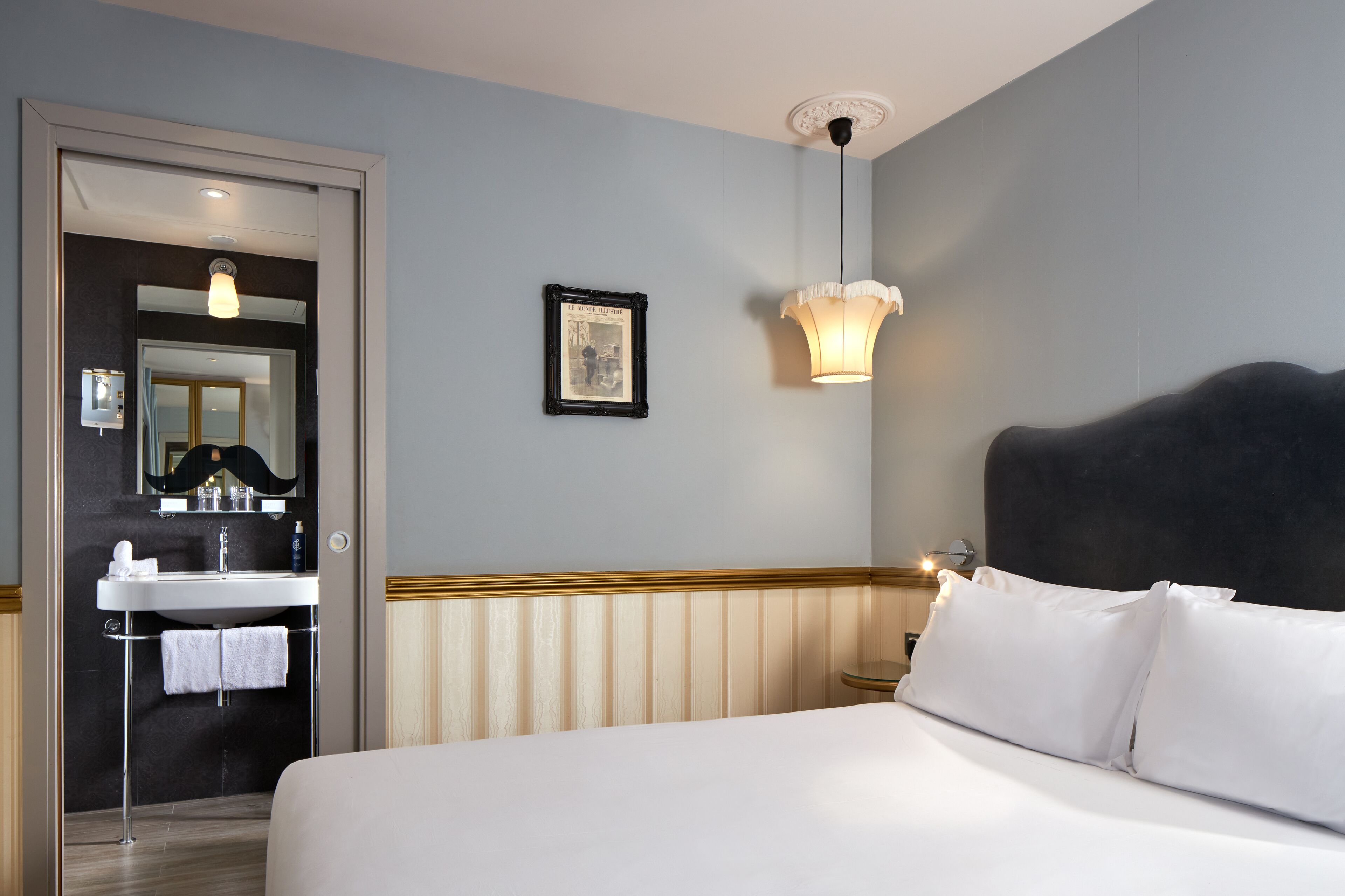 Et hotelværelse med seng, natbord, lampe på væggen, et billede i ramme, og et badeværelse med vask og håndklædestang.